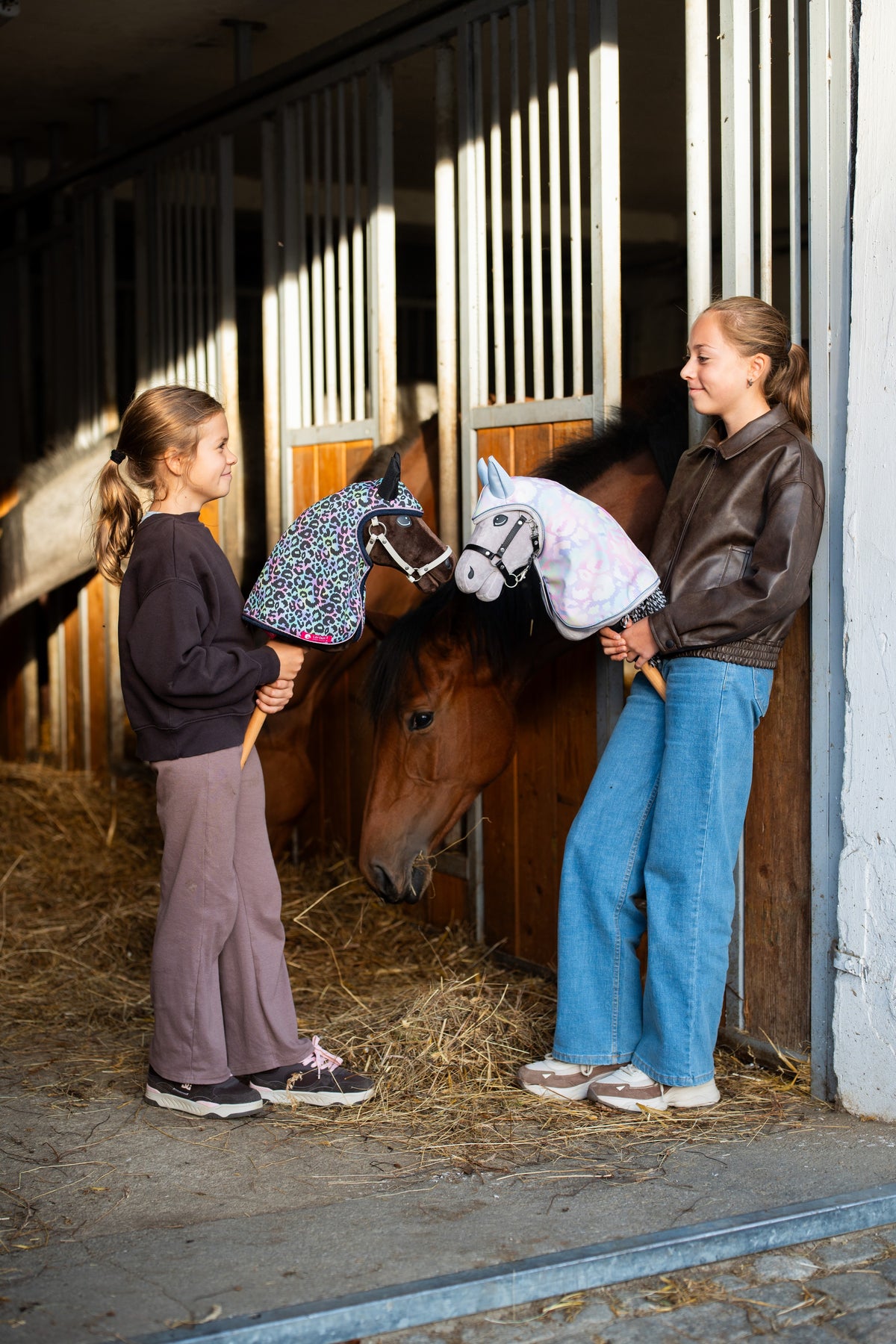 Two girls in a stable with Hobby Horse Alfredo and a grey Hobby Horse in colourful hooded raincoats by a bay horse