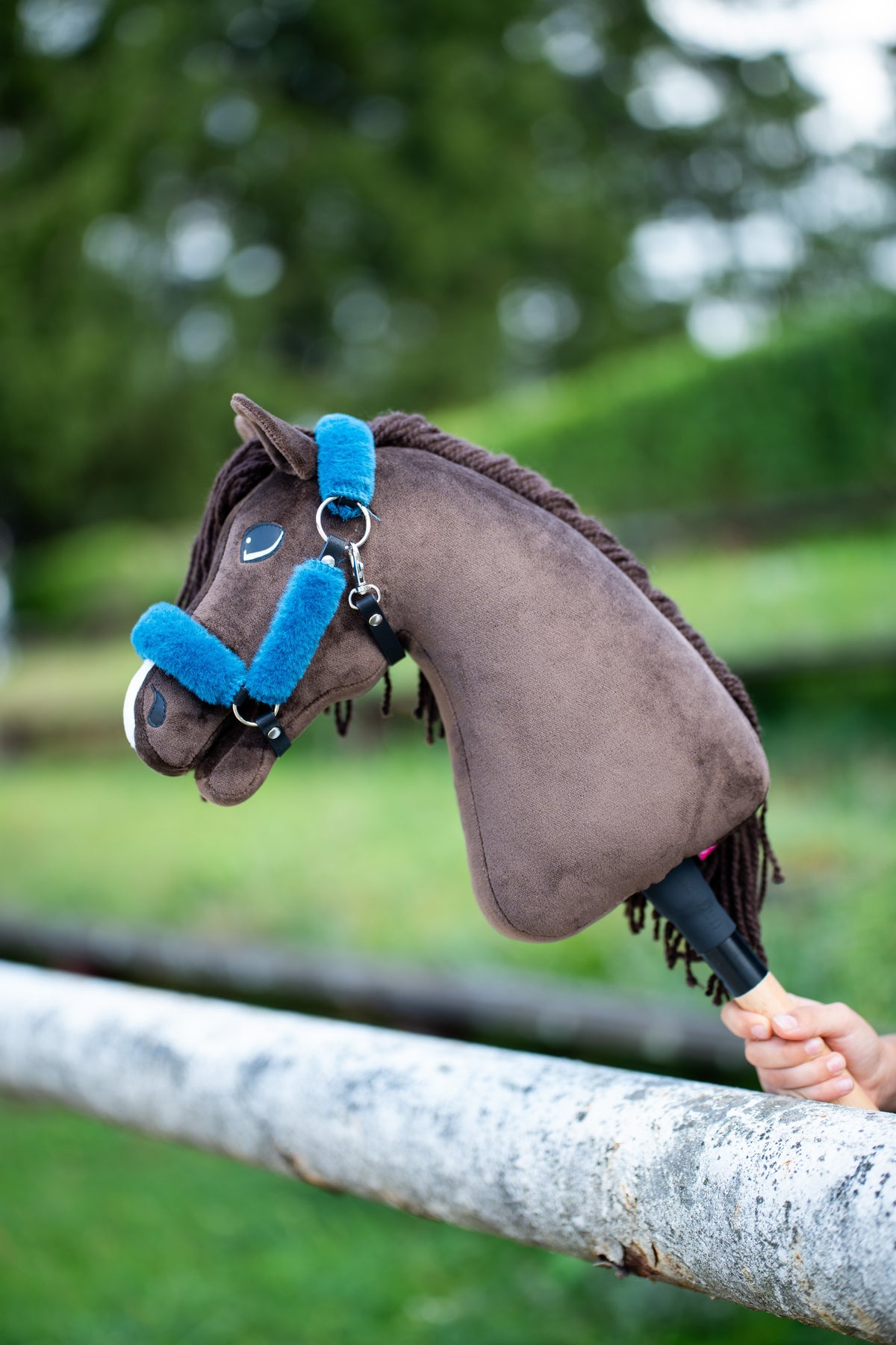 Hobby Horse Chianti in a black halter with petrol blue fur, resting on a white birch rail in front of green bushes
