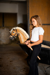 Girl in a white top standing in a stable beside a wheelbarrow, holding Hobby Horse Colby in a black halter