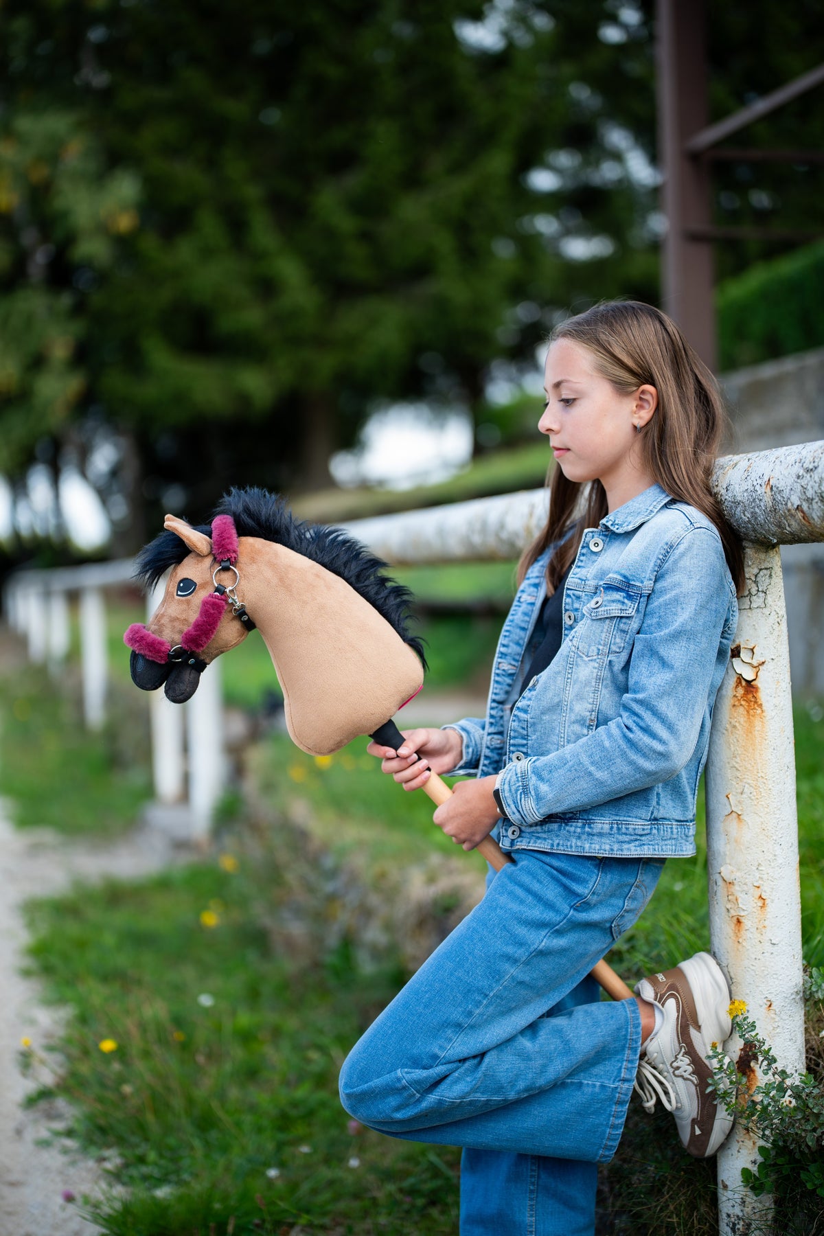Girl in a denim jacket leaning on a white rail, holding the light brown Hobby Horse in a raspberry fur halter