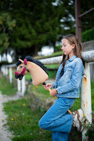 Girl in a denim jacket leaning on a white rail, holding the light brown Hobby Horse in a raspberry fur halter