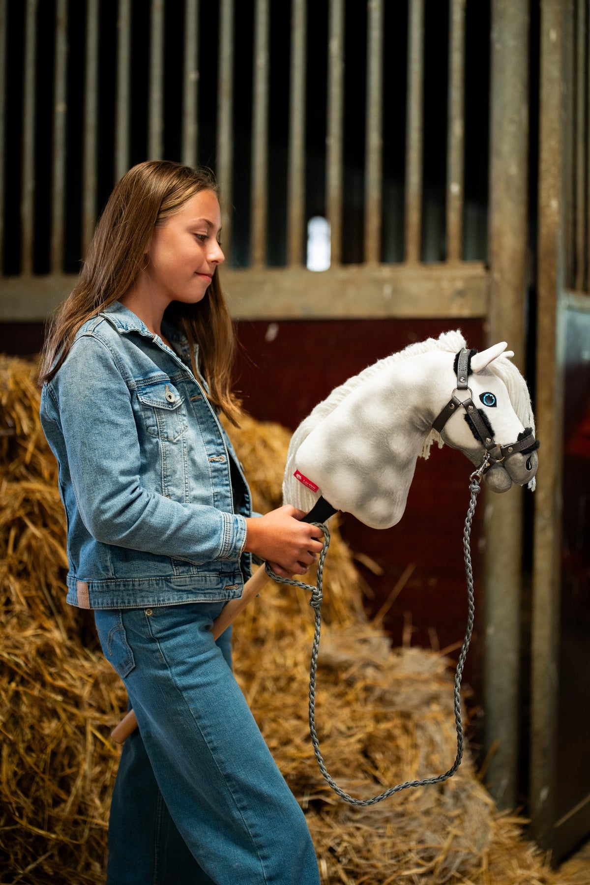 Flicka i jeansjacka vid halmbalar håller Hobby Horse Elektra, här med mörkgrå grimma med päls och flätat grått grimskaft