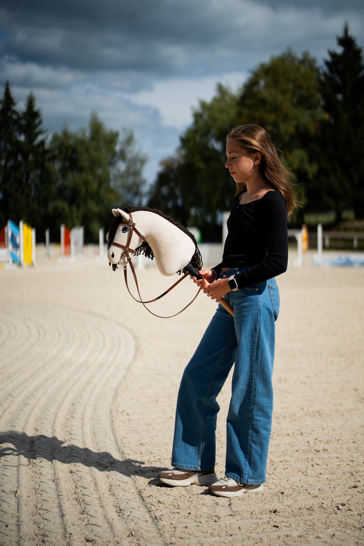 Girl on a sand arena with colourful jumps holding a cream Hobby Horse in the brown complete bridle with noseband