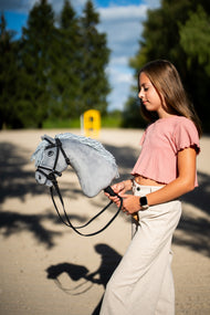Girl in a pink top walking across a sand arena with Hobby Horse Grace in a black bridle with reins