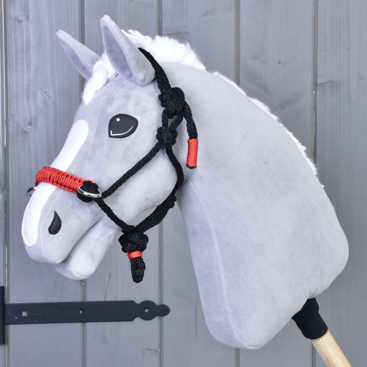 Grey plush Hobby Horse with a white mane wearing the black and red rope halter in front of a grey wooden wall