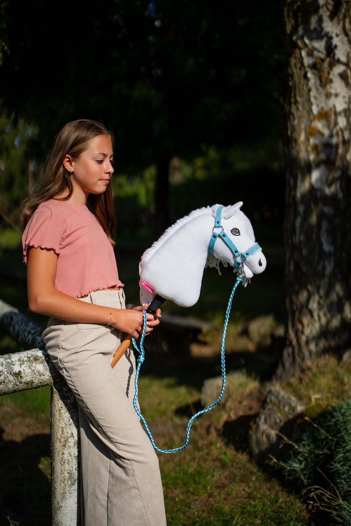Flicka i rosa topp vid en björk som håller den vita käpphästen i en ljusblå grimma med ett slingrande grimskaft