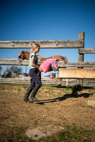 Smiling girl in riding boots walks Hobby Horse Pinkie, here in a turquoise halter, across a paddock past a chestnut horse