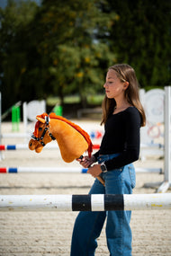 Girl in a black top on a sand arena with show jumps, holding a chestnut Hobby Horse in a black halter with white fur