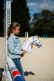 A girl in a denim jacket leans on a jump stand in a sand arena, holding a white Hobby Horse in the grey halter with mint fur