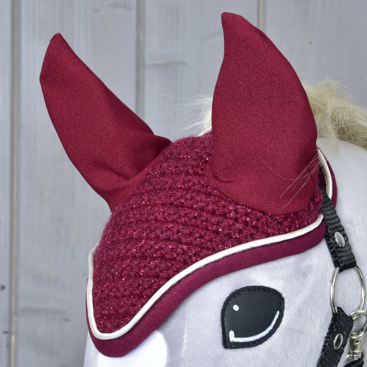 Close-up of the Ear bonnet crocheted Lux Maroon on a grey Hobby Horse: wine red ears, sparkly crochet and white piping