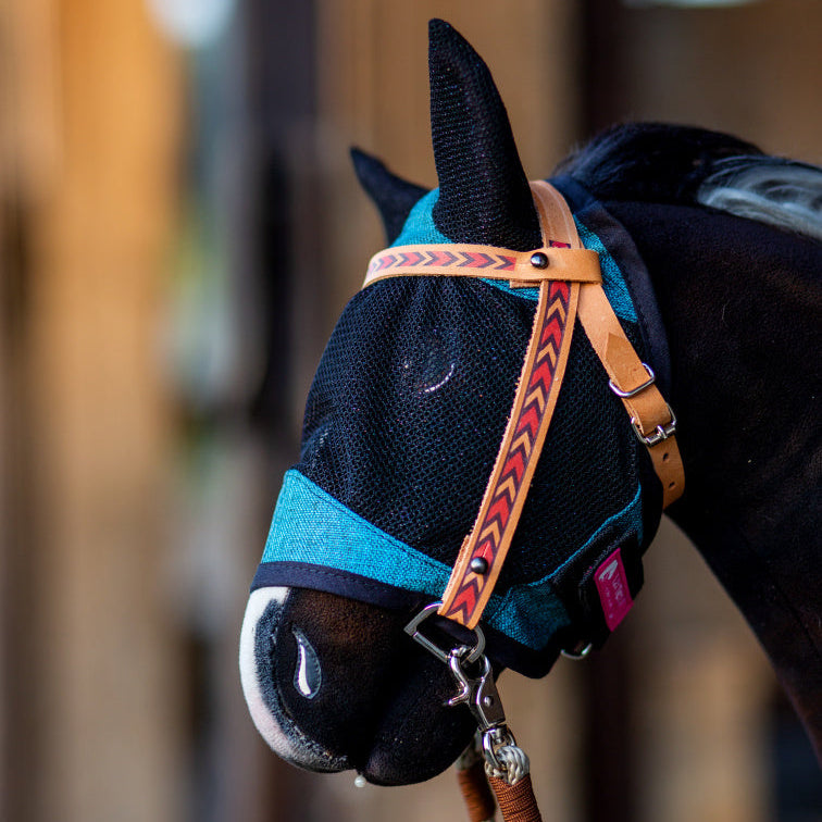 Närbild av en käpphäst med en blå flugmask i nät med öronskydd och en brun grimma med rött pilmönster, utrustning för Hobby Horsing. Bakgrunden är mjukt suddig.