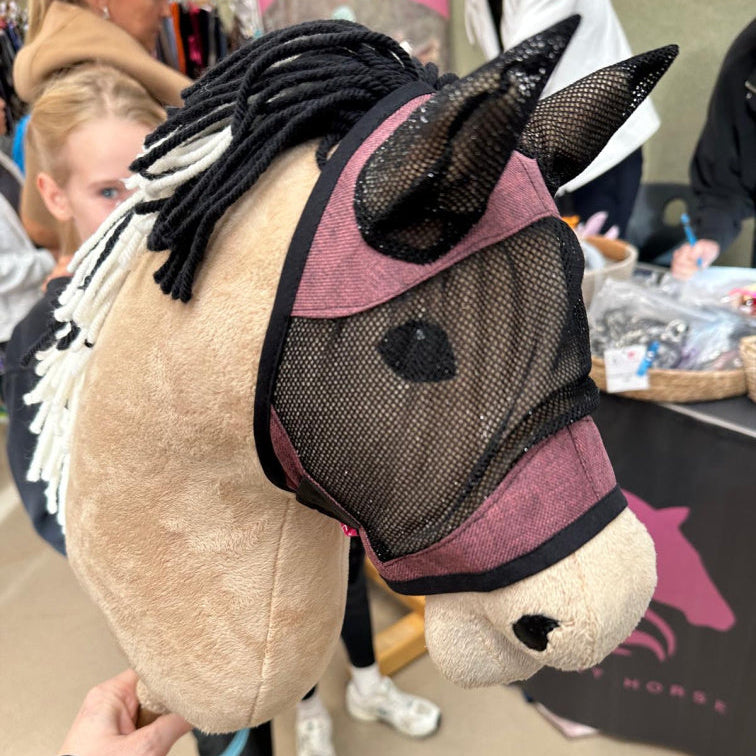 Närbild av en handgjord käpphäst med nätmask och svartvit man av garn. I den suddiga bakgrunden syns människor och ett stånd med hästprodukter, som visar den kreativa andan i Hobby Horsing.
