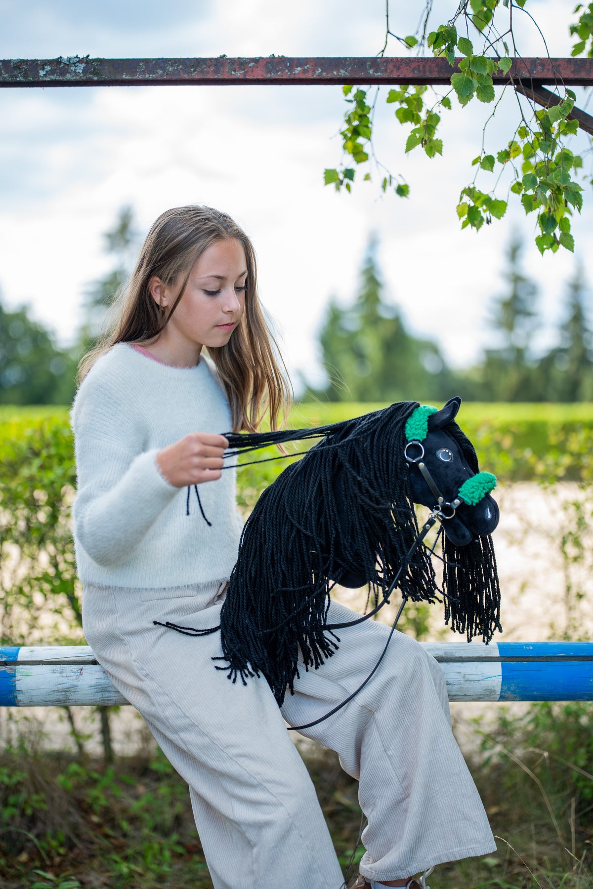 Flicka i vit tröja som sitter på en hinderbom och samlar ihop tyglarna på den svarta käpphästen i en grön grimma med päls