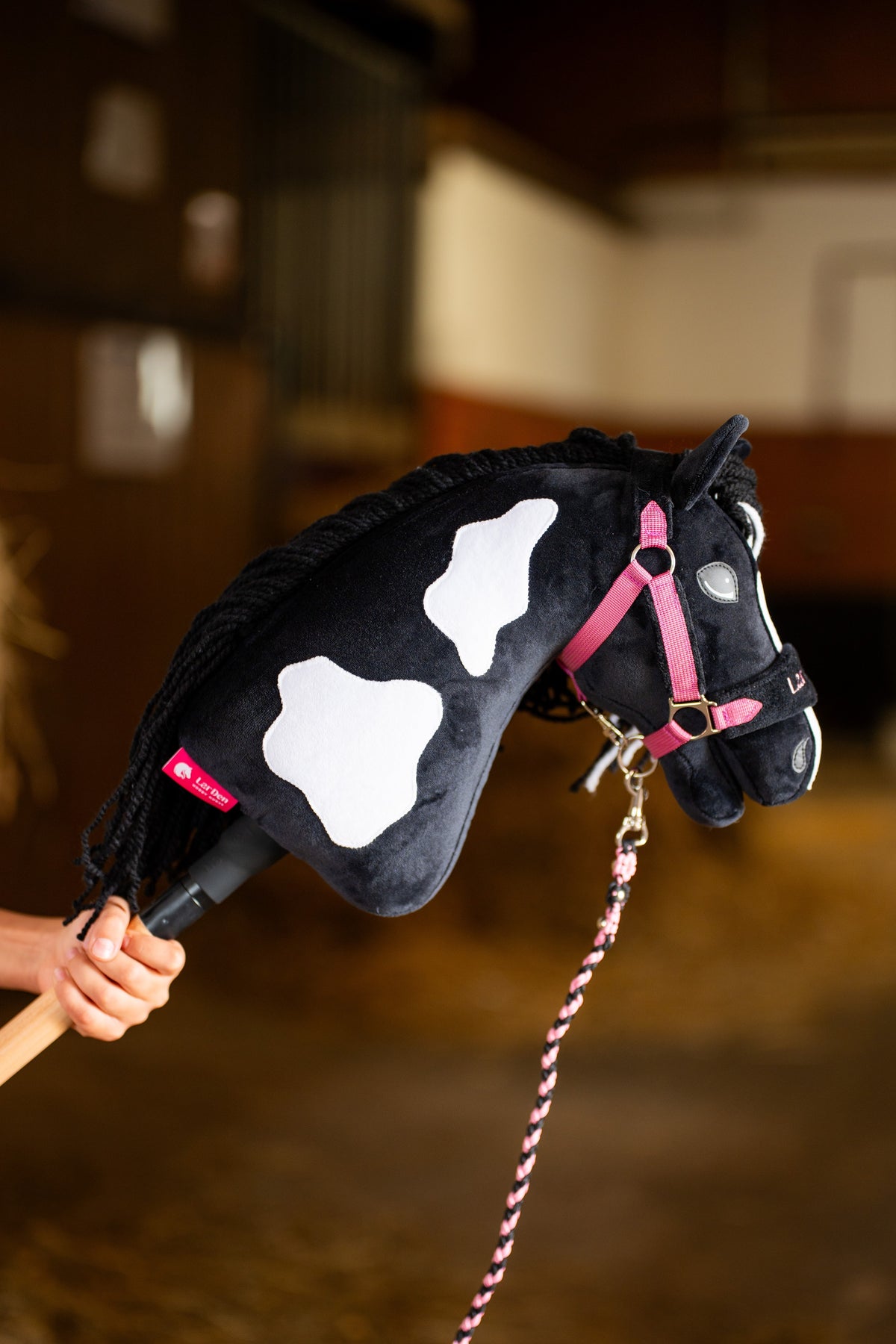 Hobby Horse Merlin med stora vita fläckar i rosa grimma av vävt band och ett rosa och svart flätat grimskaft, i ett stall