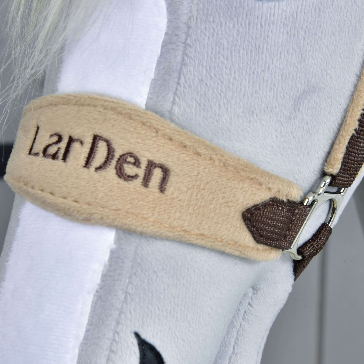 Närbild av nosremmen i beige fleece med LarDen broderat i mörkbrun tråd, bredvid en vit bläs