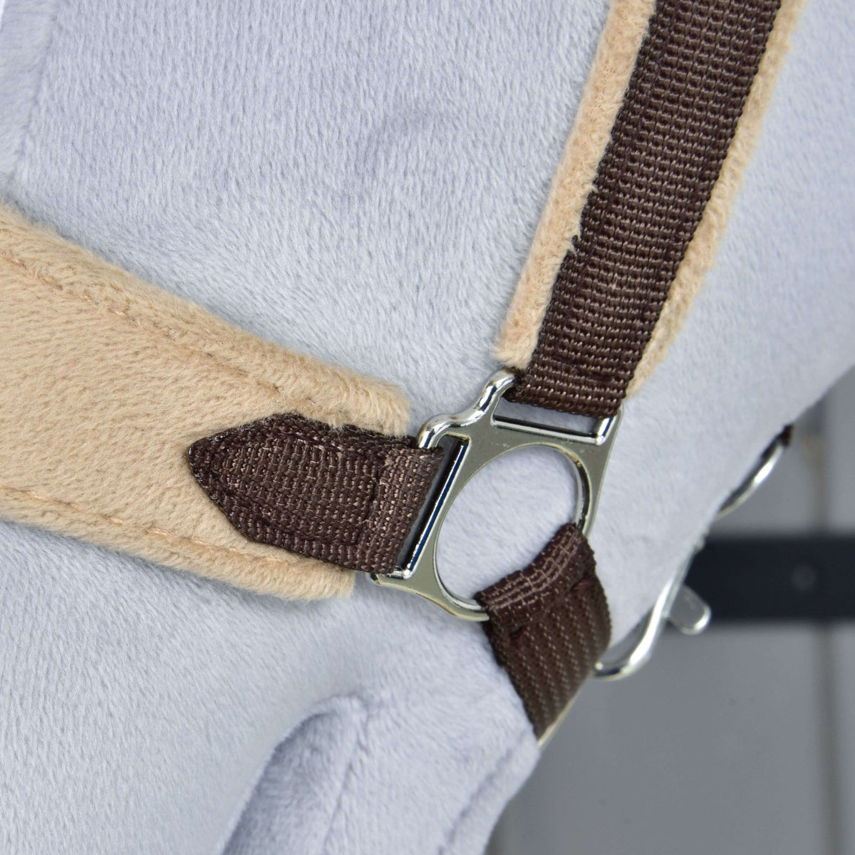 Närbild av den silverfärgade sidoringen som förenar den mörkbruna kindremmen med nosremmen i beige fleece på den bruna grimman med päls