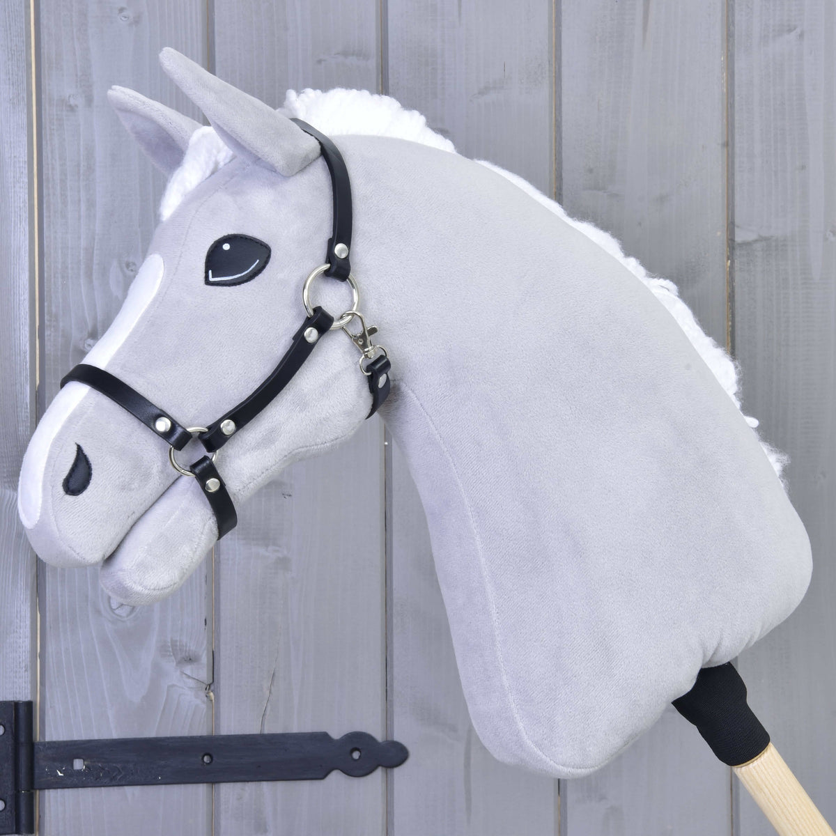 Grey plush Hobby Horse with a fluffy white mane and blaze wearing the black leatherette halter in front of a grey wooden wall