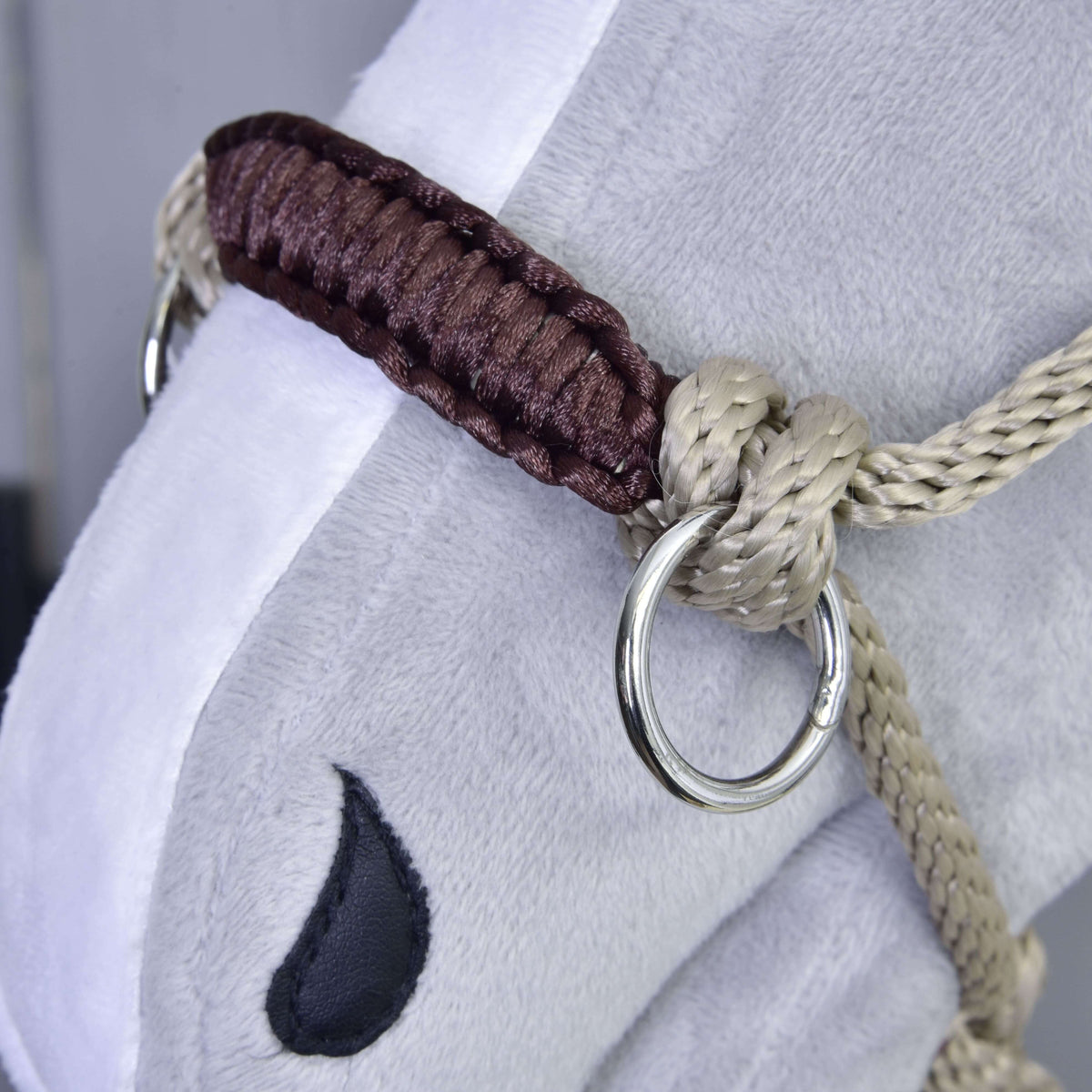 Närbild på den beige repgrimman med brun flätad nosrem och silverring på en grå käpphäst i plysch