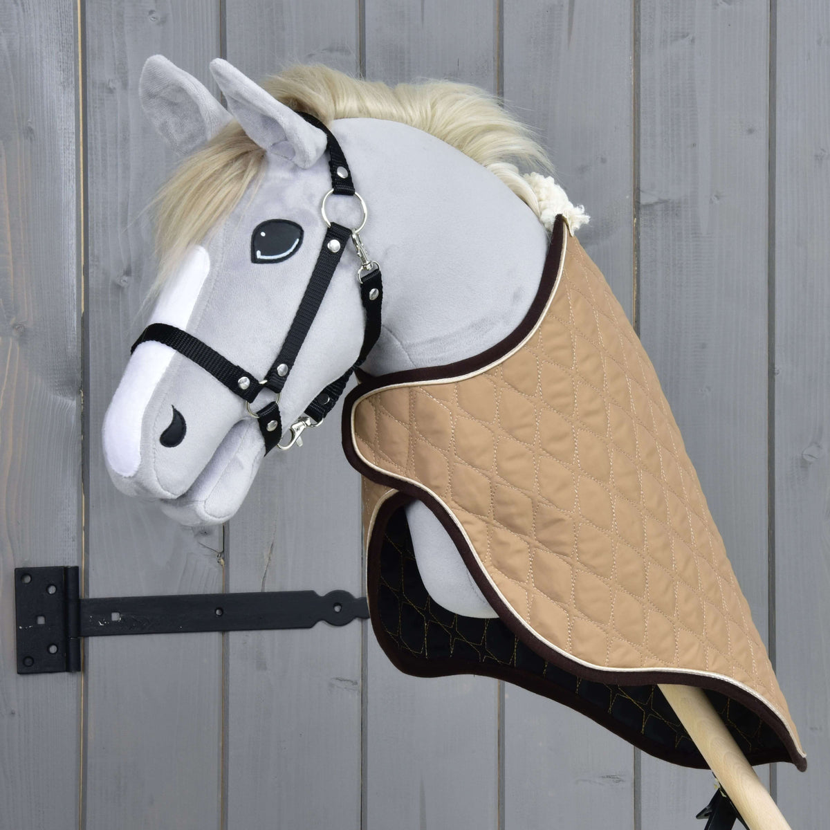 Grey plush Hobby Horse in a black halter wearing the beige quilted travel rug, front three-quarter view by a grey wooden wall