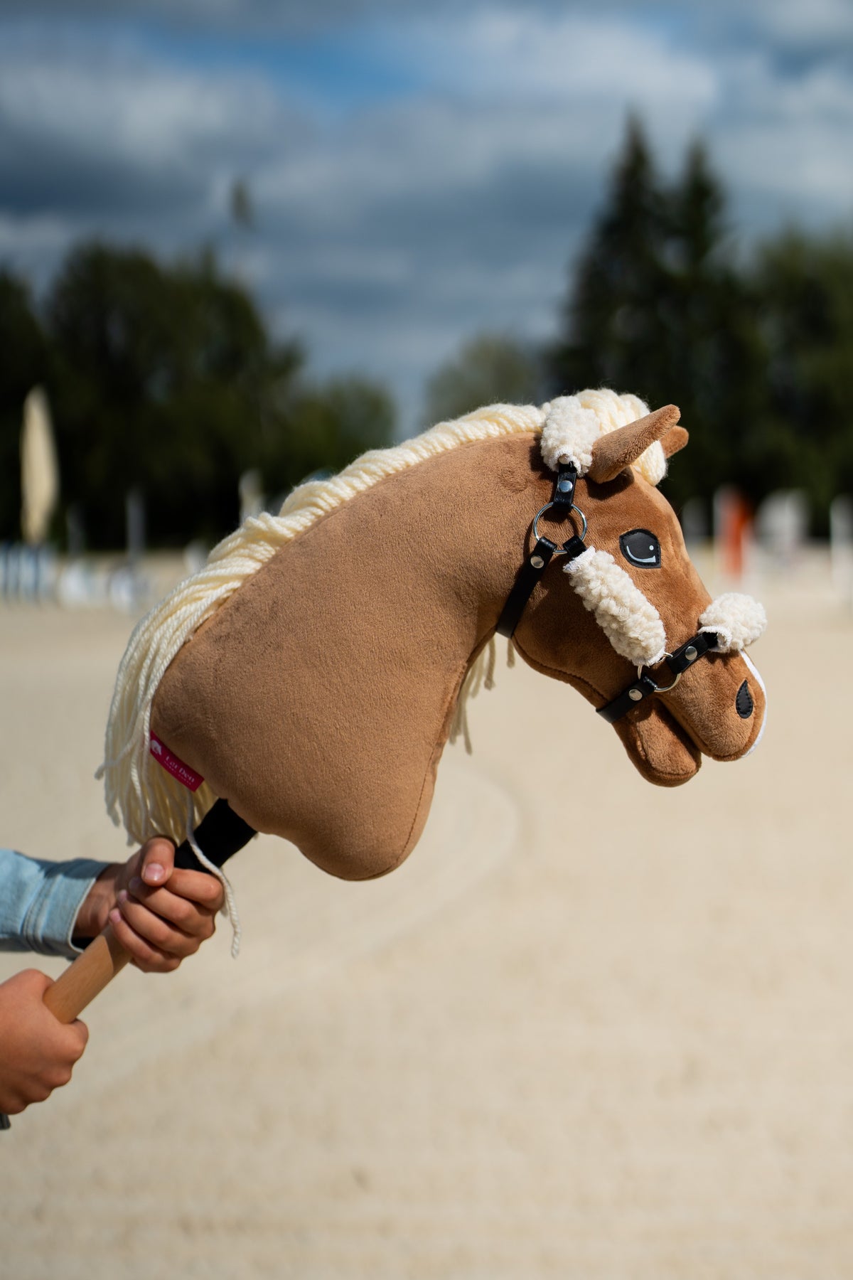 Ljusbrun Hobby Horse Smart i den svarta grimman helt klädd med krämvit päls, upphållen på en sandbana under en molnig himmel