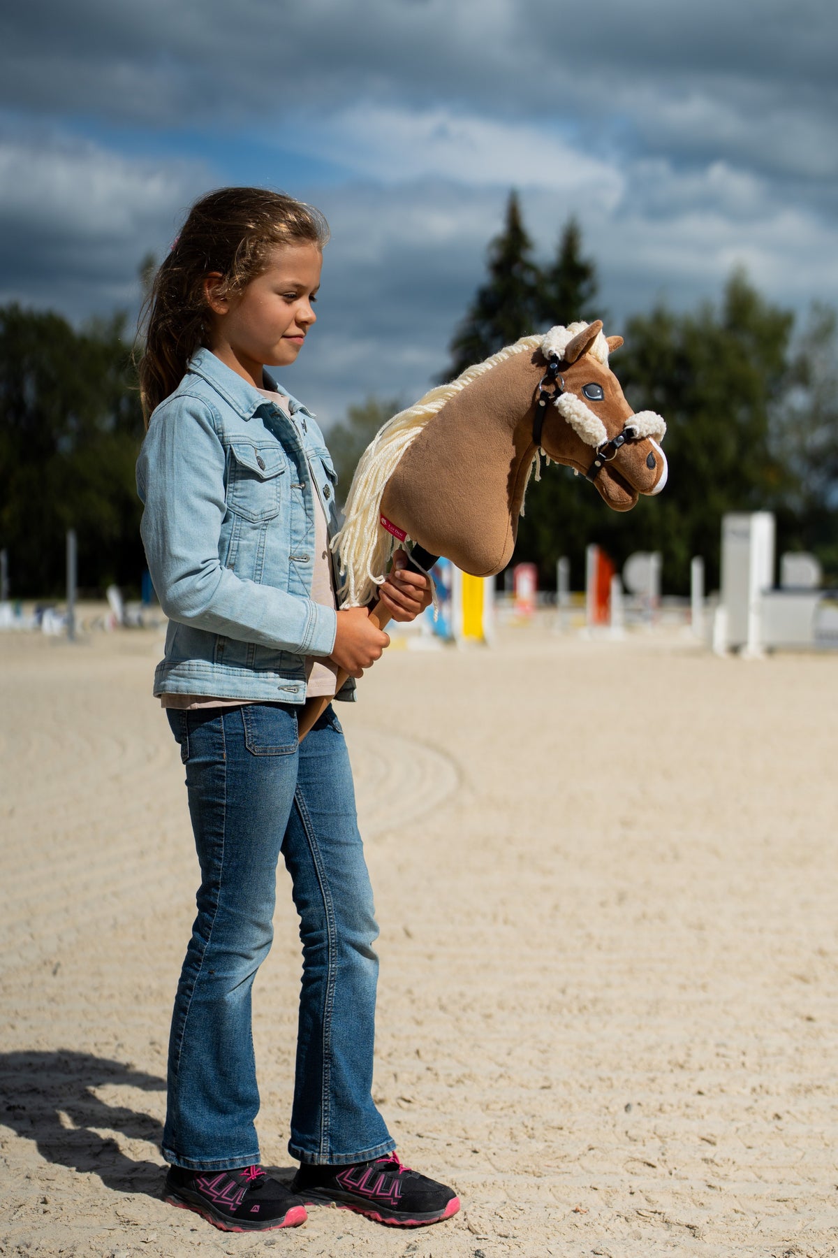 Flicka i jeansjacka på en sandbana med hinder, med en ljusbrun käpphäst i grimman med gräddvit päls