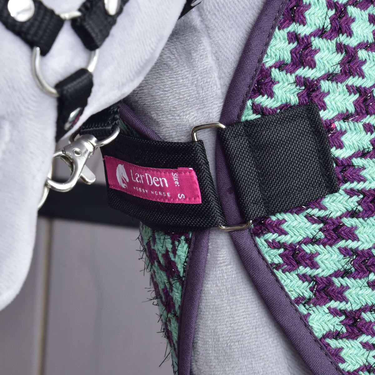 Bröstremmen på Hobby Horse stalltäcke grönt stängd med D-ringar över ett grått plyschbröst, rosa etikett från LarDen syns