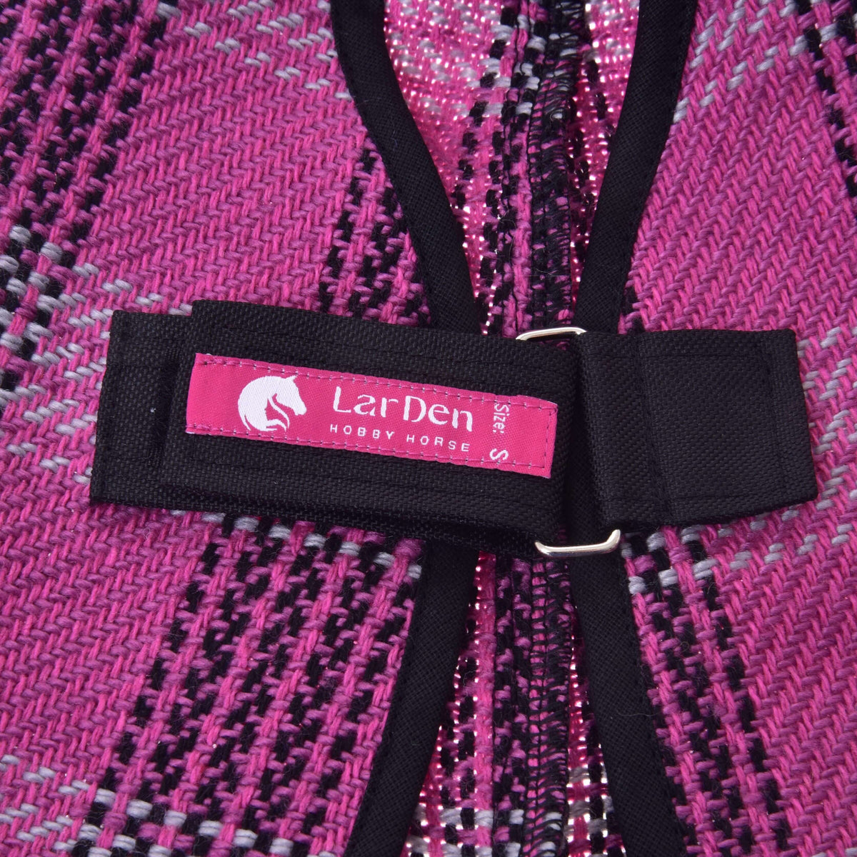 Närbild på den rosa etiketten från LarDen på den svarta bröstremmen på Hobby Horse stalltäcke rosa, mot det vävda rutmönstret