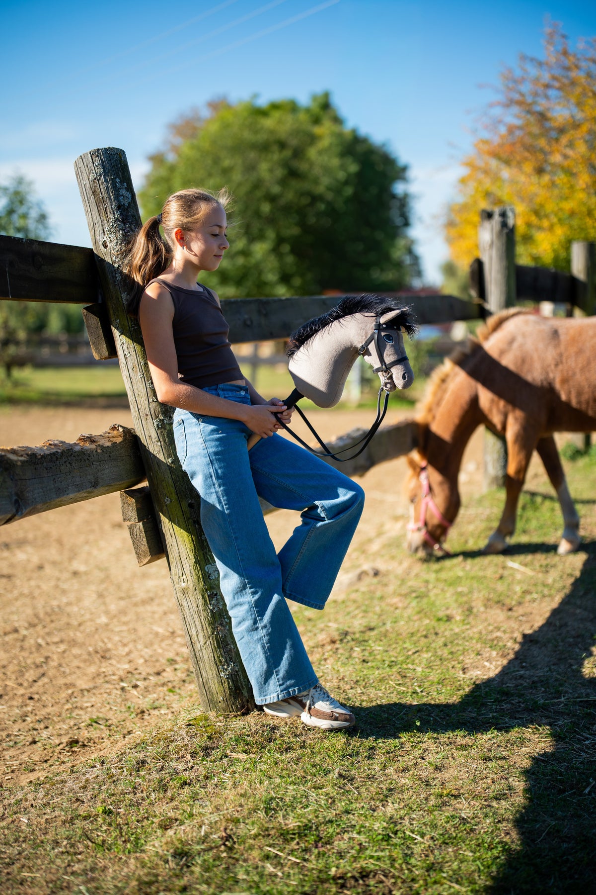Flicka lutad mot ett hagstaket i höstsol med Hobby Horse Thor i svart träns, en fuxfärgad ponny som betar bakom