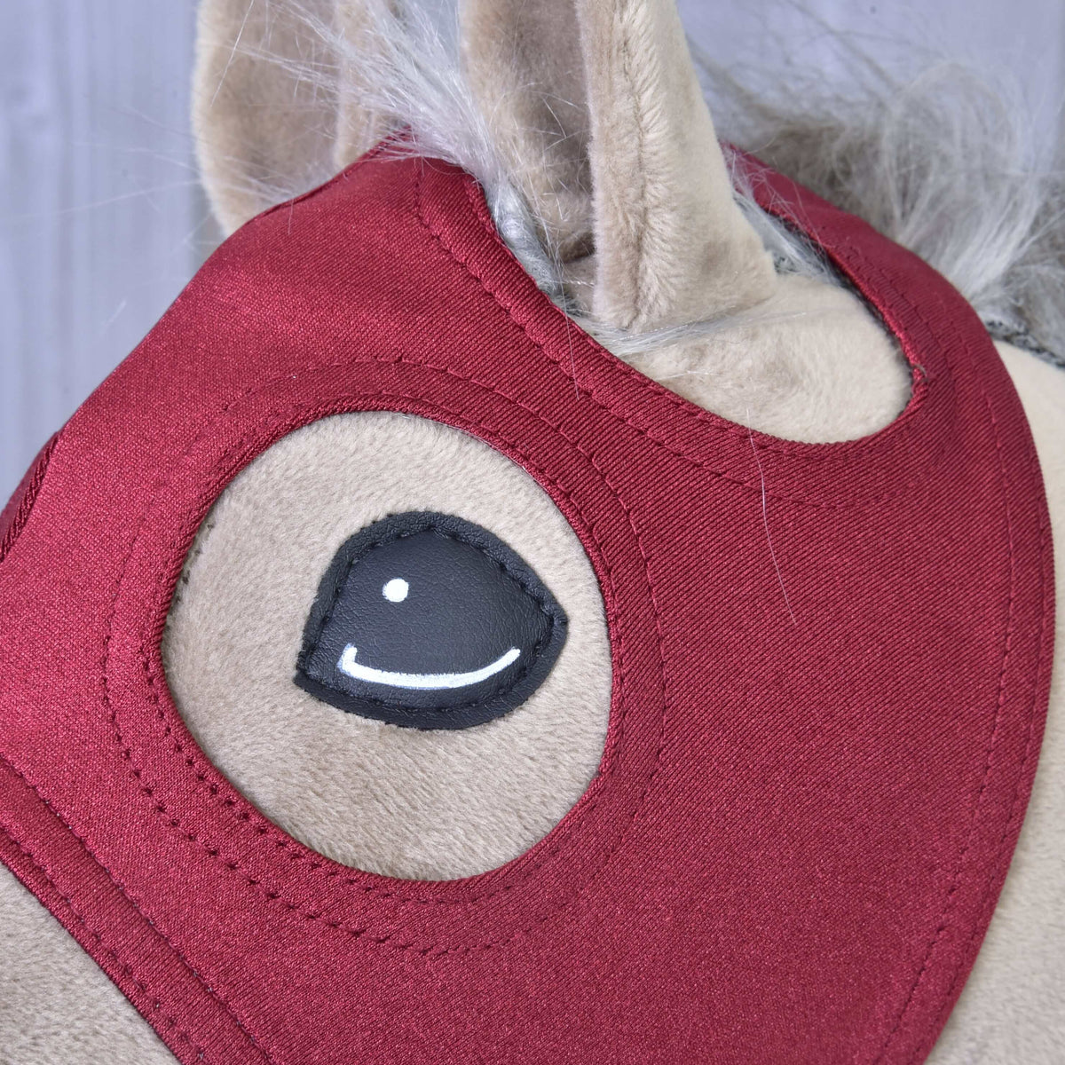 Eye and ear of a beige Hobby Horse framed by the burgundy mask, with double stitching around the openings and a greyish mane