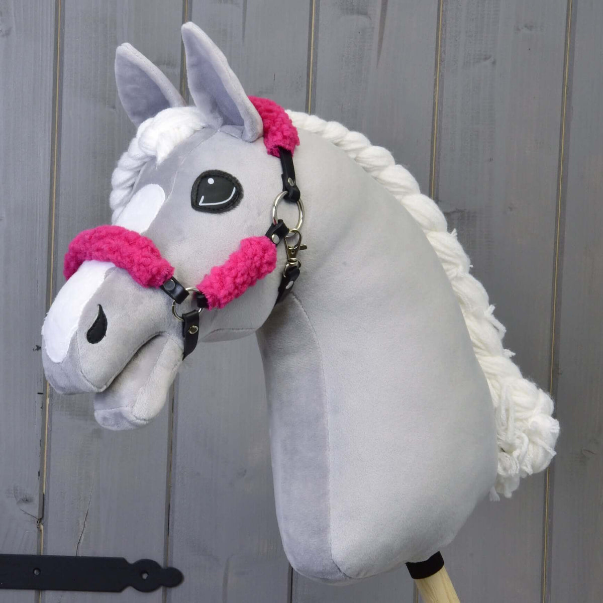 Grey plush Hobby Horse with a braided white mane wearing the black halter with complete pink fur, grey wooden wall