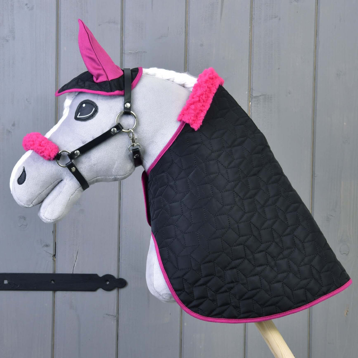 Grå käpphäst i plysch med det svarta quiltade täcket, flughuvan och grimman med fuchsiafärgad päls, trävägg